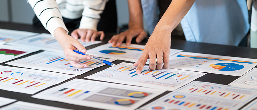 Hände mehrerer Teammitglieder zeigen auf bunte Geschäftsdiagramme und Berichte während eines Meetings – symbolisiert gemeinsame Analyse von Qualitätskosten und strategische Maßnahmenplanung zur Effizienzsteigerung.